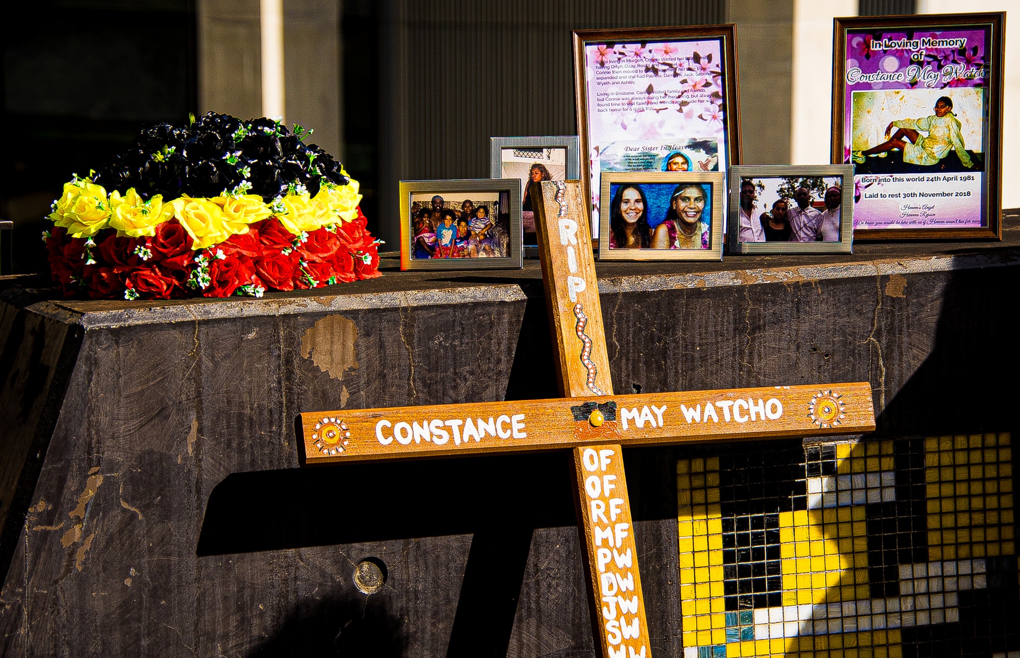 A photograph of the memorial for Constance May Watcho. There is a flower arrangement of black, yellow and red roses, a cross with Ms Watcho's name and designs painted on it, and a cluster of framed photographs of Ms Watcho and her family.