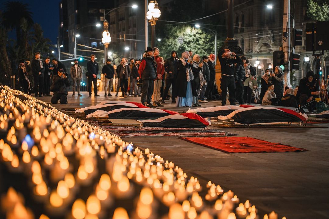 vigil for palestine on the parliament steps. Hundreds of tealight candles on the steps. Palestinian flag covering fake dead bodies on the ground. A group of people standing around. 