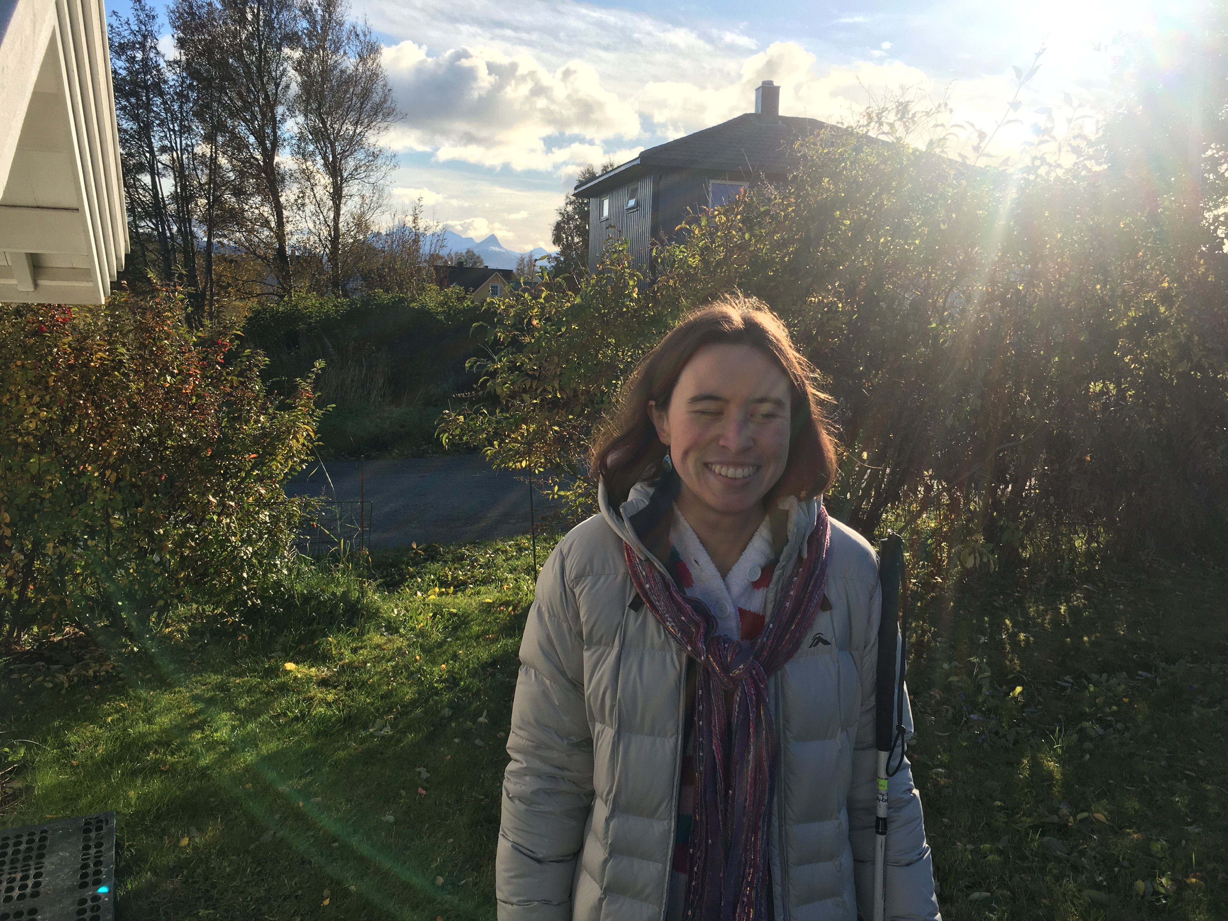 Image description: A white non-binary person with shoulder length hair stands in winter greenery outdoors, smiling. They're holding their white cane, and mountains are visible behind them.