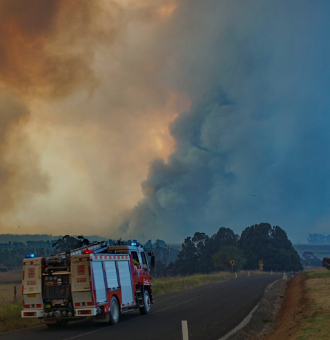 A CFA truck drives towards a plume of bushfire smoke along a rural road