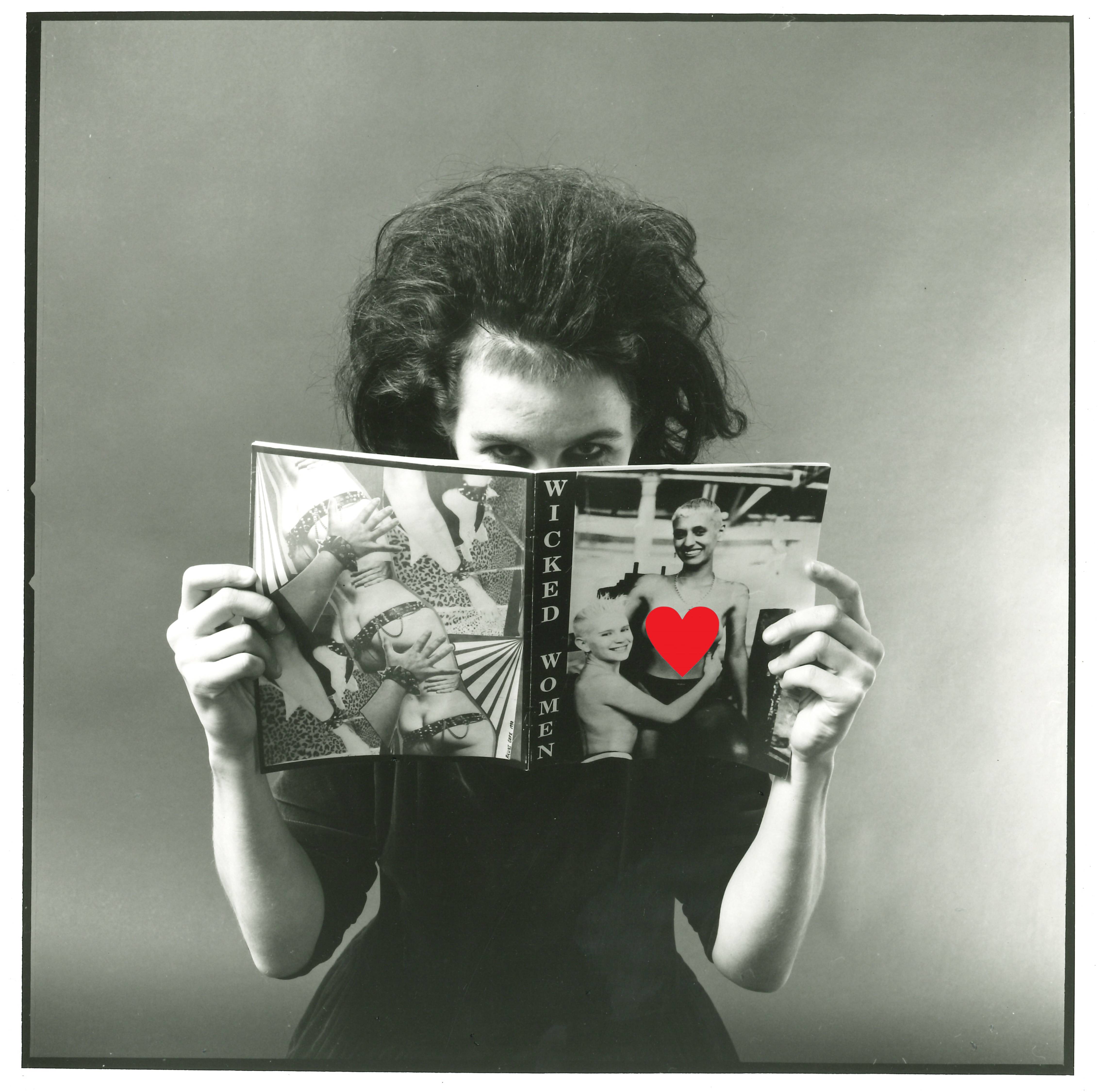 Black and white portrait style image showing a woman with teased hair and black eyeliner in a black velvet dress reading a copy of Wicked Women magazine. She holds the magazine in front of her face with just her eyes looking out at us, disrupting the relationship between viewer and viewed