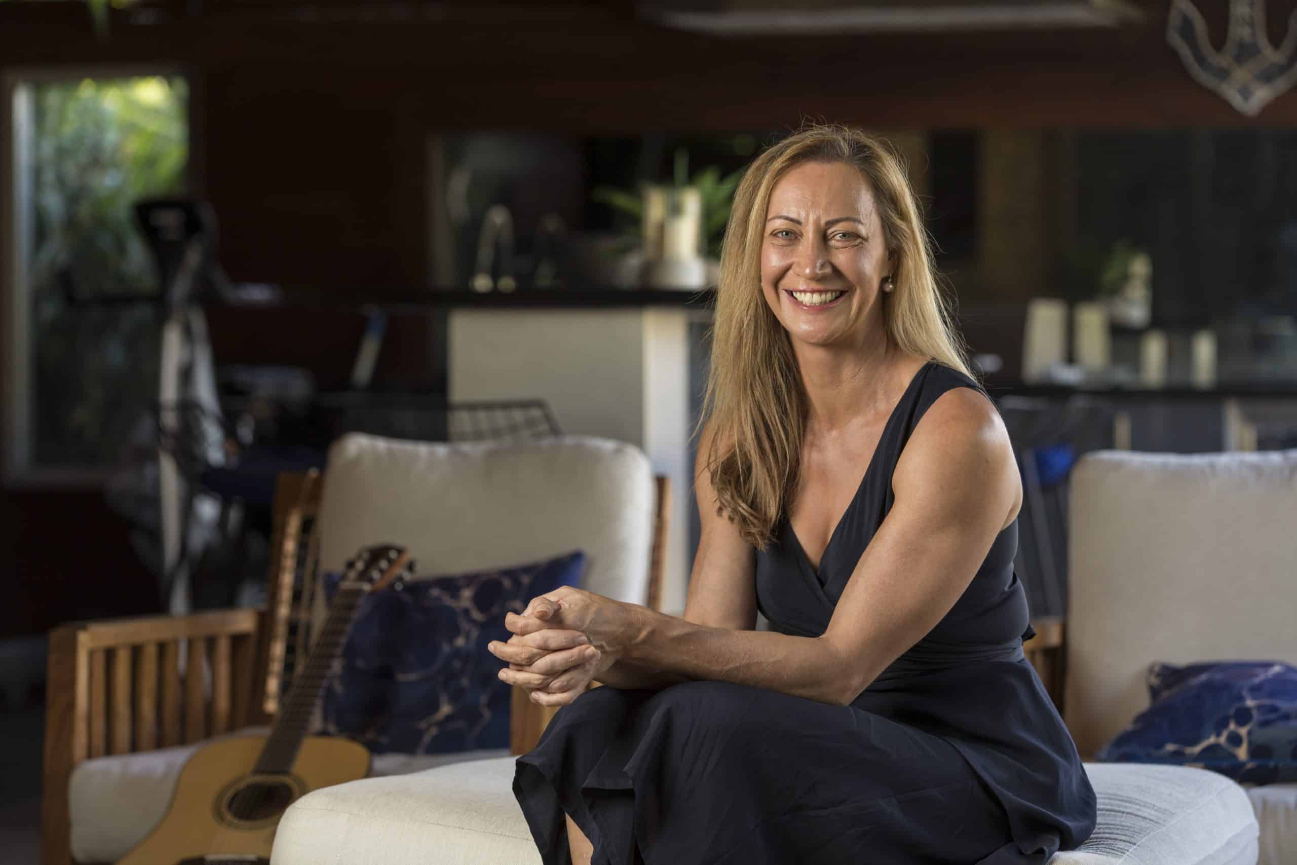 A photograph of Dr Tracy Westerman sitting on a light beige ottoman seat. She is facing the camera and smiling, with her elbows resting in her lap and hands clasped.