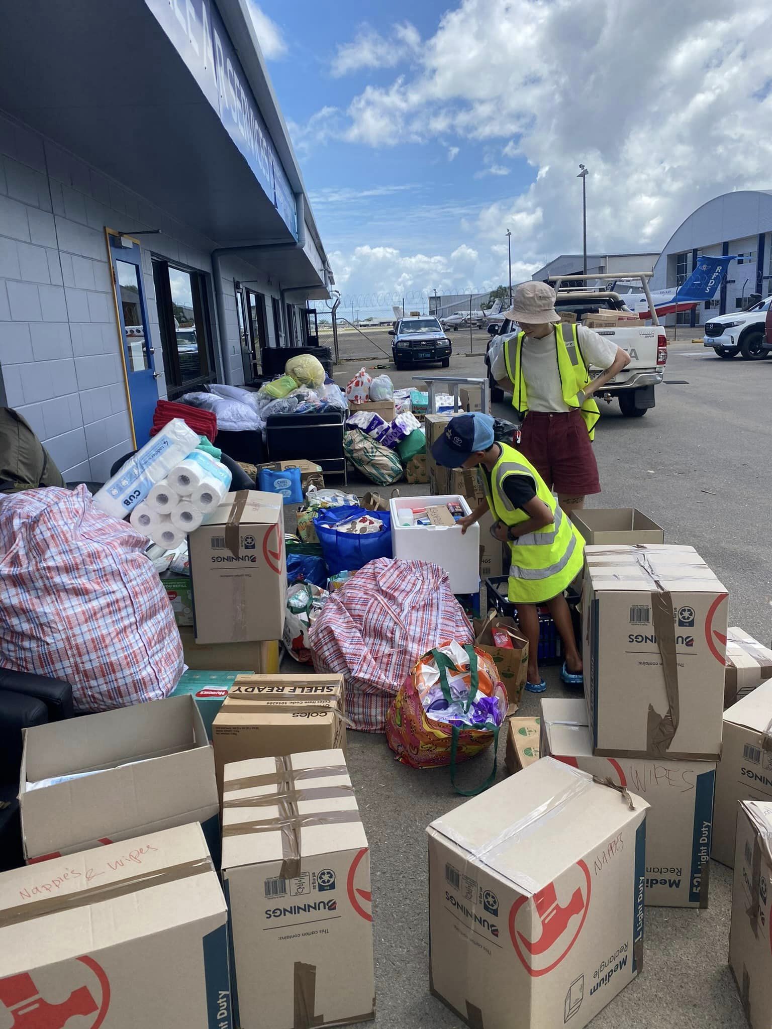 Two people wearing yellow fluro vests work with boxes and bags of supplies on the tarmac at Cairns airport