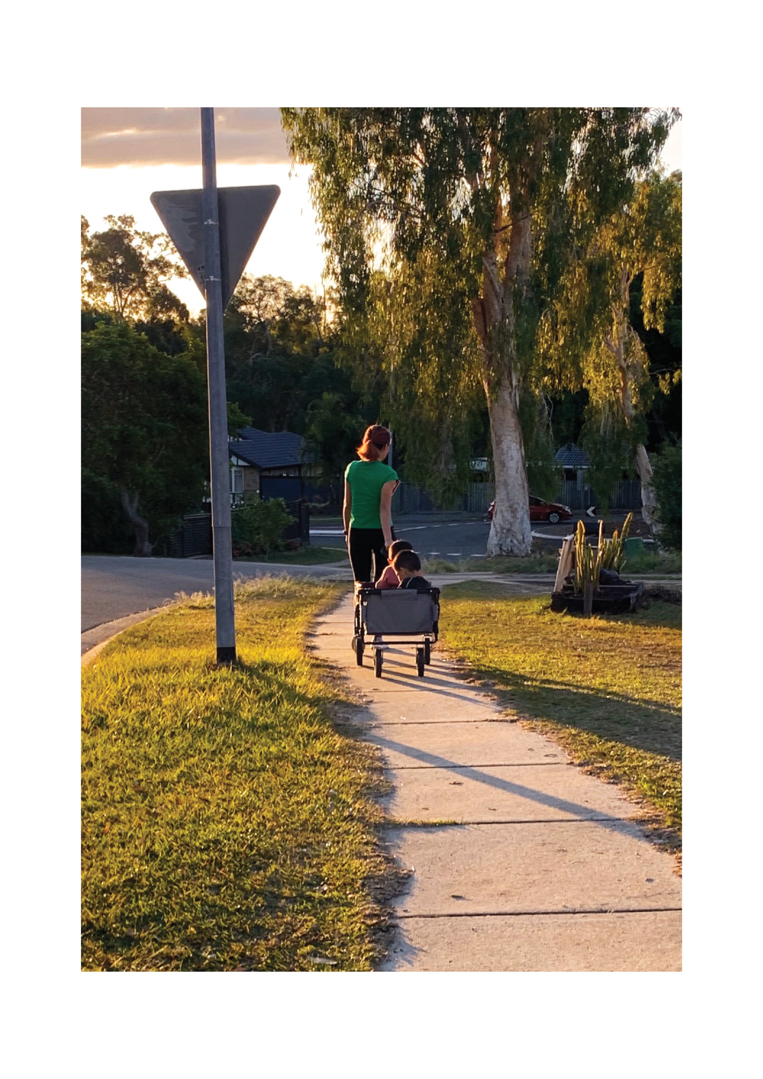 A photo of a woman pulling a trolley with two young children inside. They are walking on the sidewalk away from the camera. There's a road sign on the left and some trees on the right.