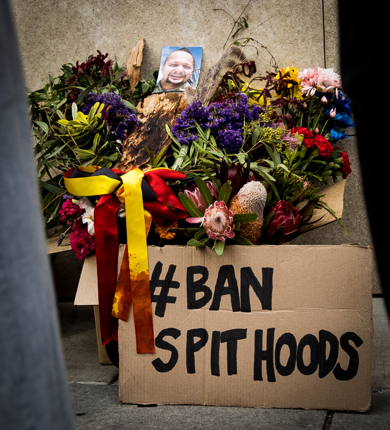 Ban Spithoods placard sits against a box of flowers, with a picture of Wayne Fella Morrison