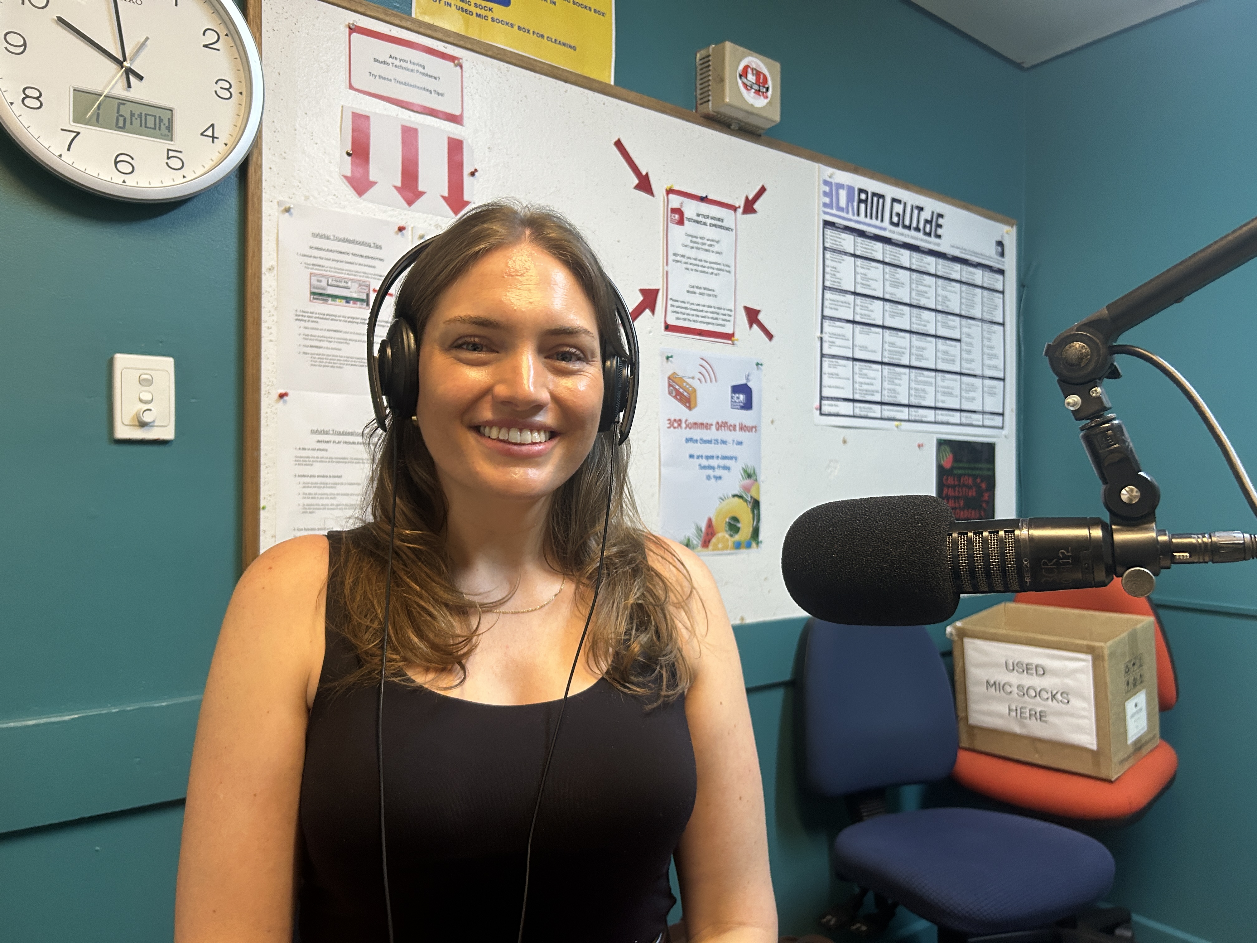 A woman sits facing the camera and smiling. She has long brown hair and is wearing headphones and on her left is a microphone. 