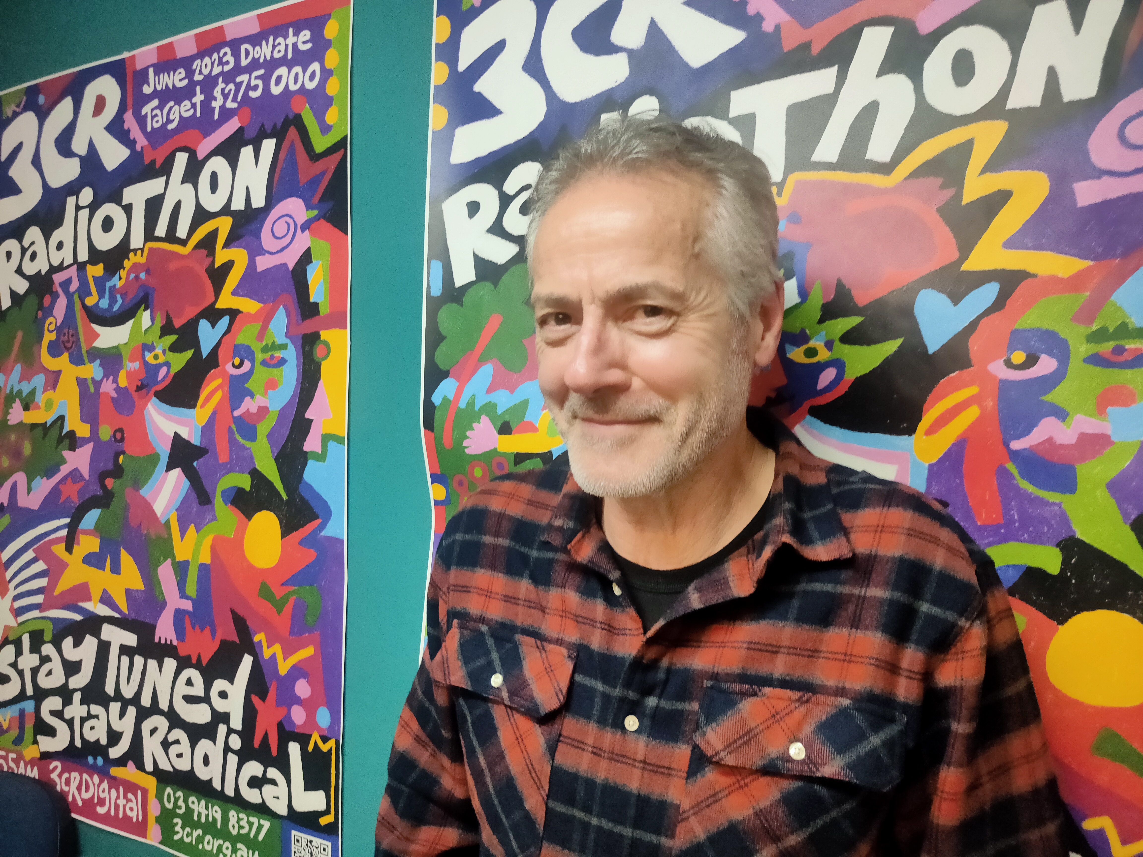 A man with greying hair wears a red and black check shirt. He looks to the side smiling