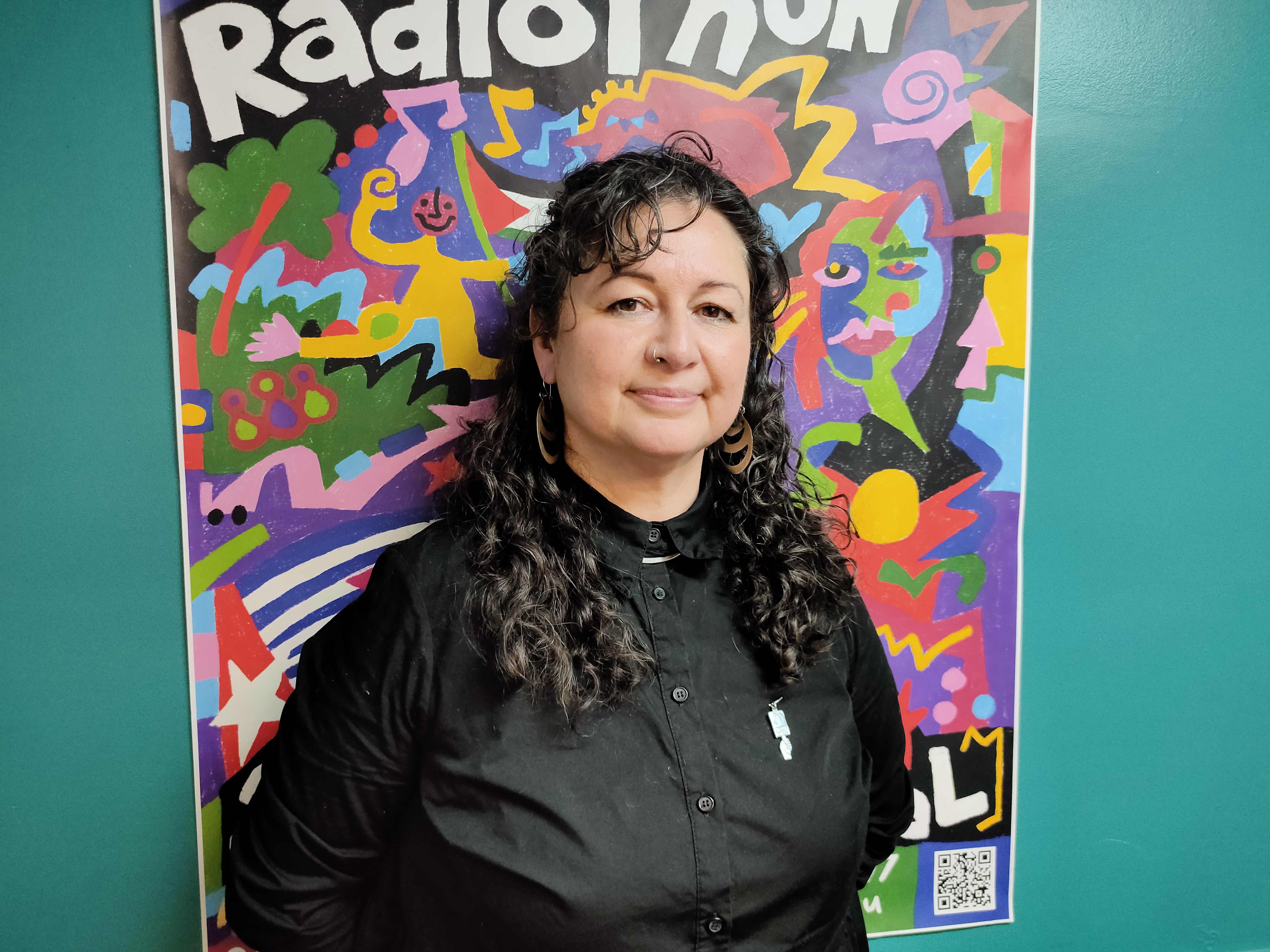 A woman with dark long curly hair and wearing a dark jumper looks sideways towards the viewer. She stands in front of a colourful poster