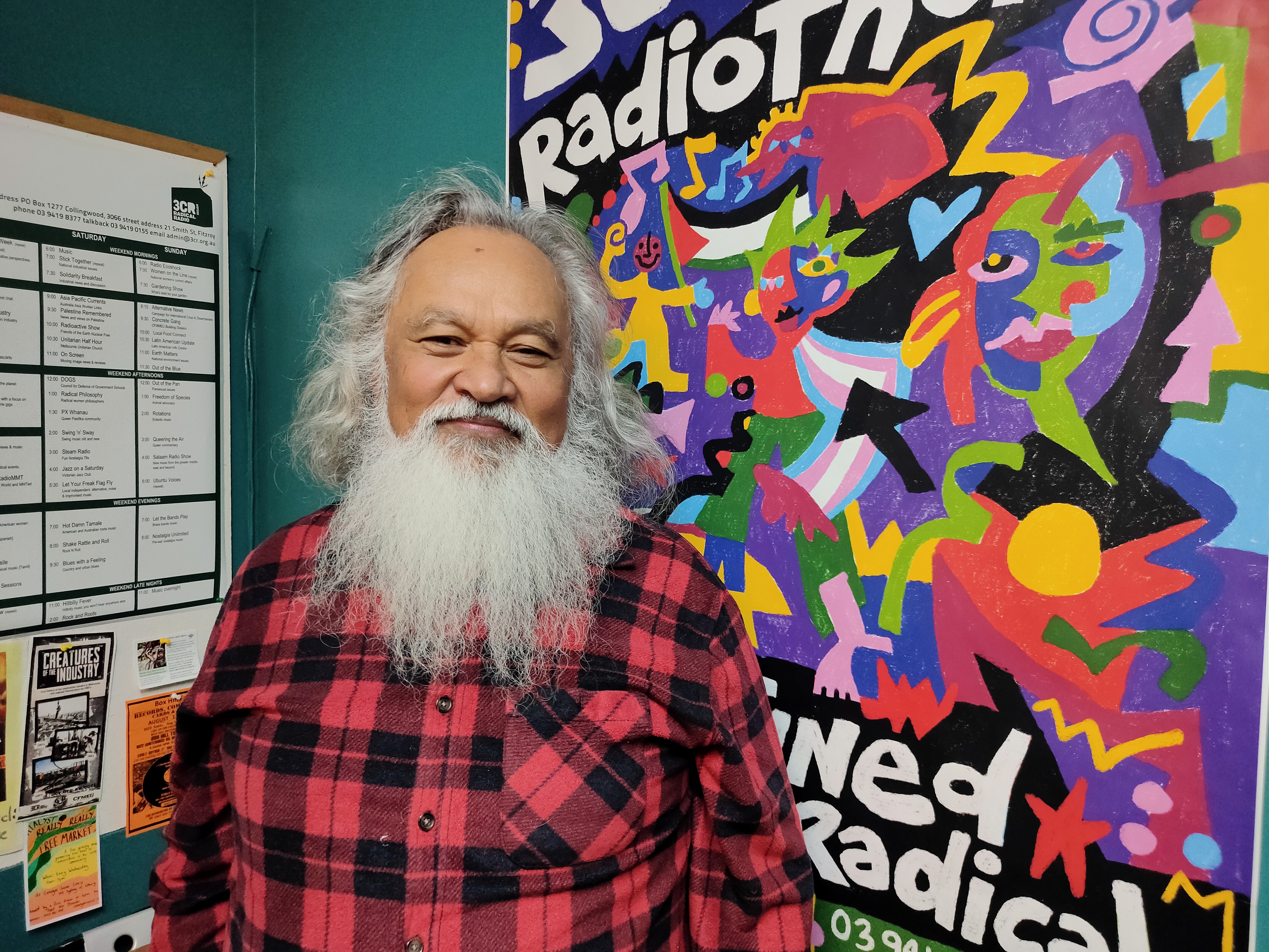 Robert stands in the 3CR studio with his red and black check shirt looking at the viewer. Behind him hangs the 2023 3CR Radiothon poster in all its technicolour glory