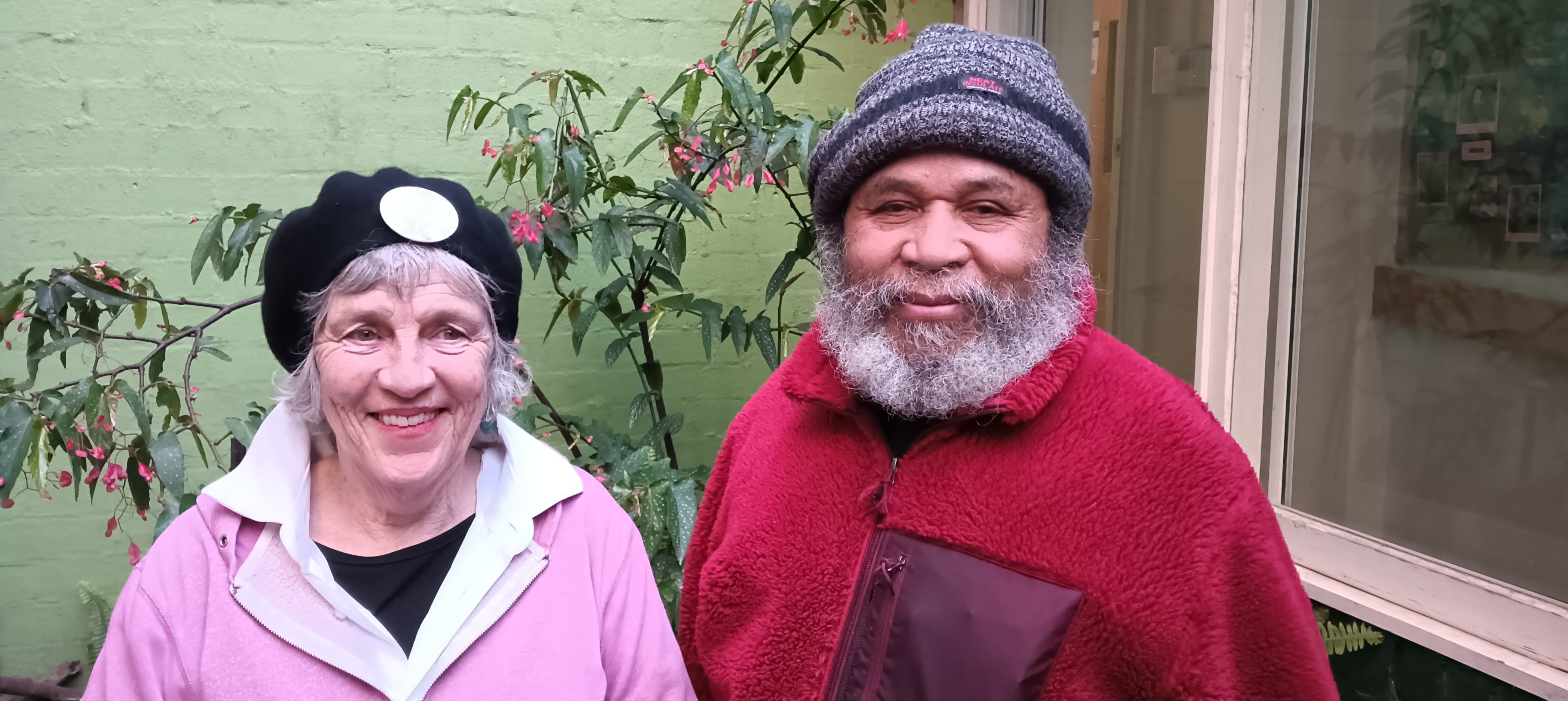 Jacob and Louise stand in the 3CR courtyard. They are wearing their winter woolies in pink and red, complete with beret and beanie.