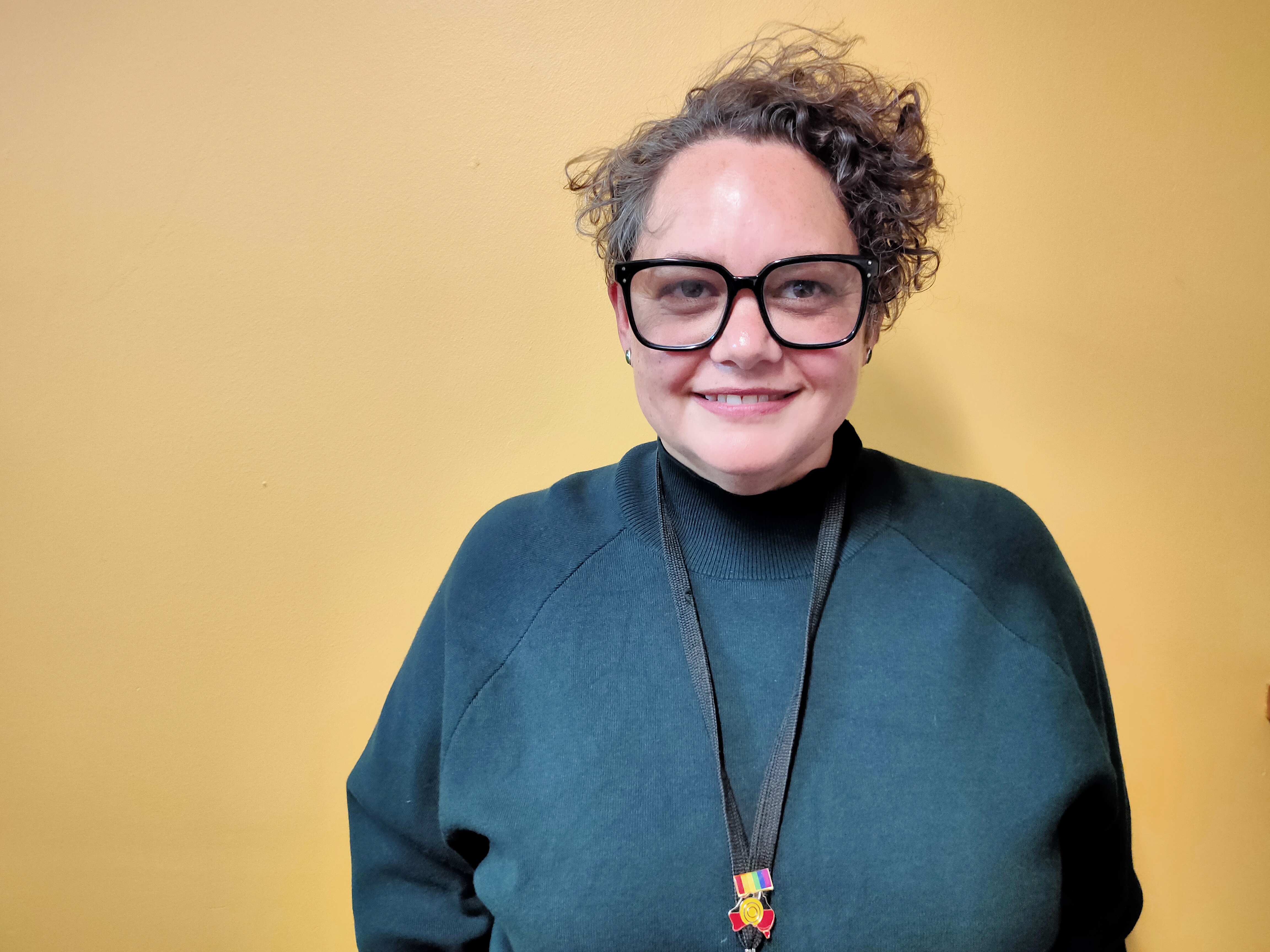 A woman in her forties looks into the camera. She wears a green jumper and black glasses. She has short curly hair