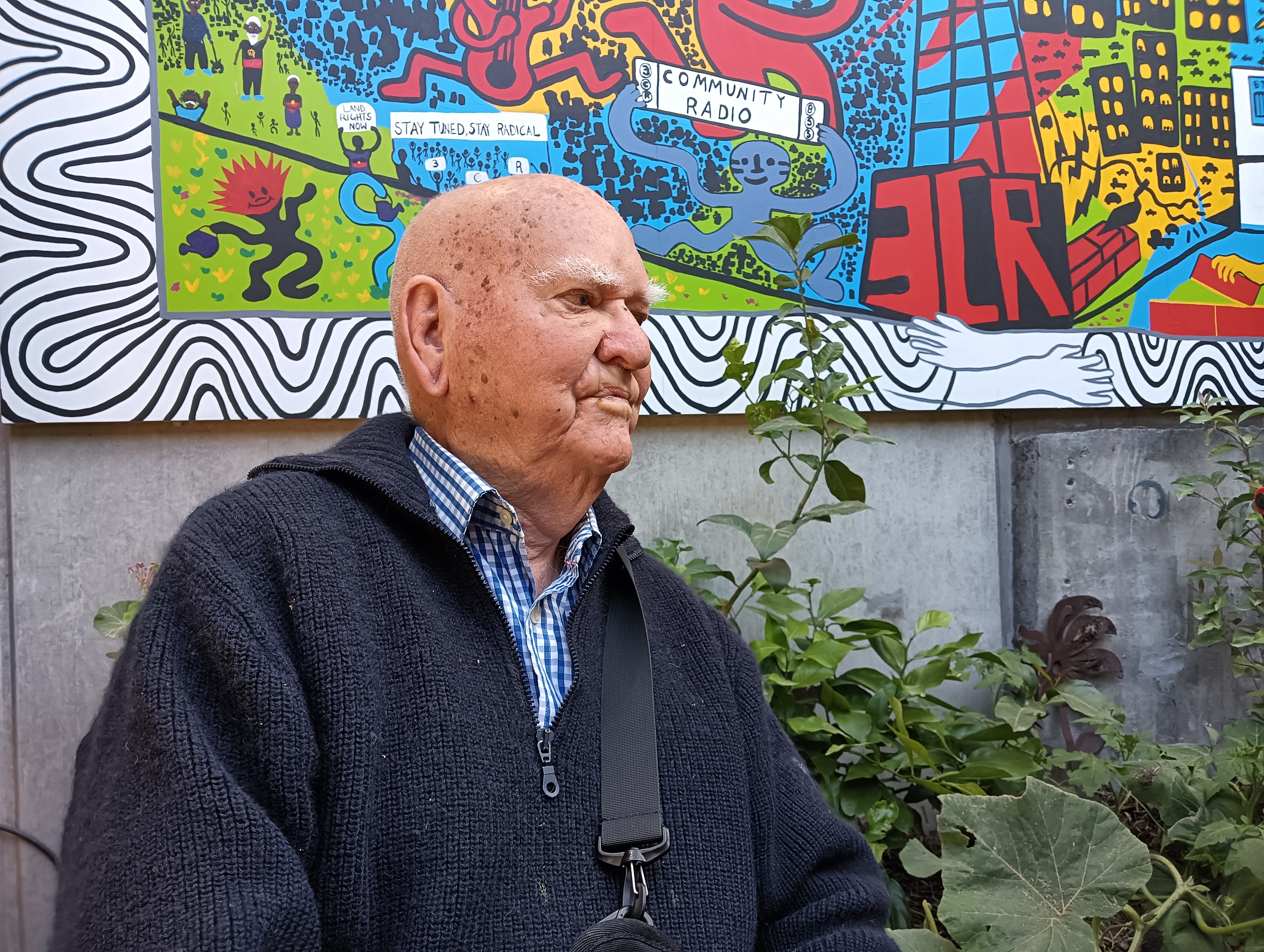 Percy sits in the 3CR courtyard and the colourful mural is behind him. He wears a blue, knitted jumper.