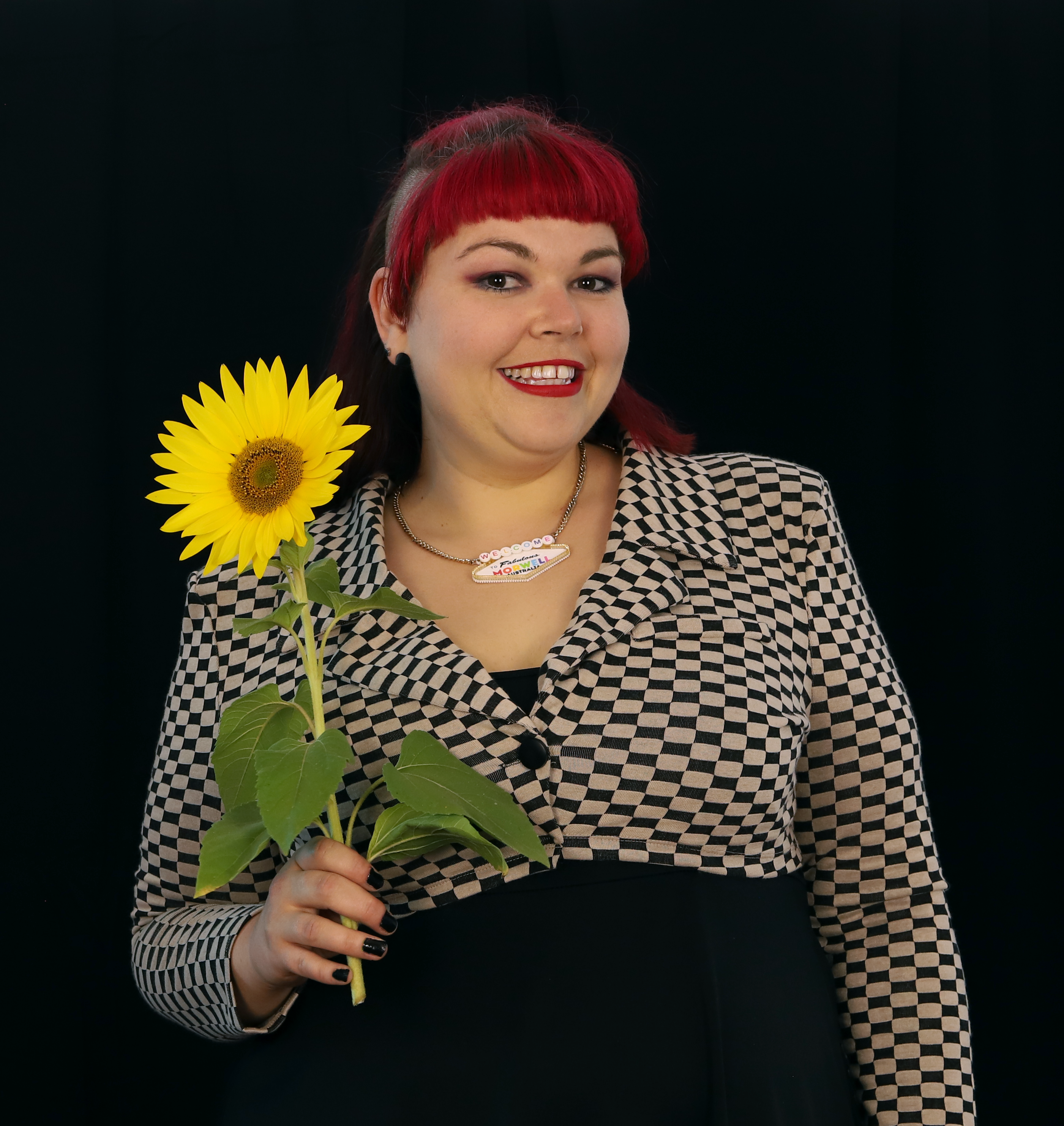 A woman stands straight on and looks into the camera holding a large sunflower up to her shoulder