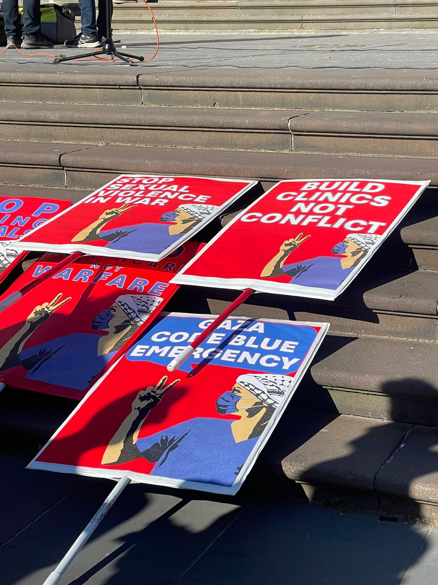 Red and blue protest signs at read "Build Clinics Not Conflict", "Gaza Code Blue Emergency"; "Stop Sexual Violence in War"