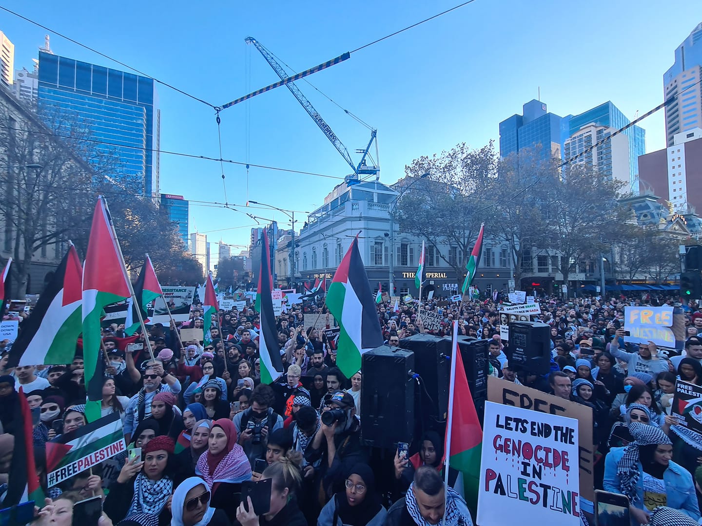 A photograph of the Spring Street and Bourke Street intersection in Narrm/Melbourne filled with people attending the Nakba Rally. Many are holding Palestinian flags and signs of solidarity with Palestine.