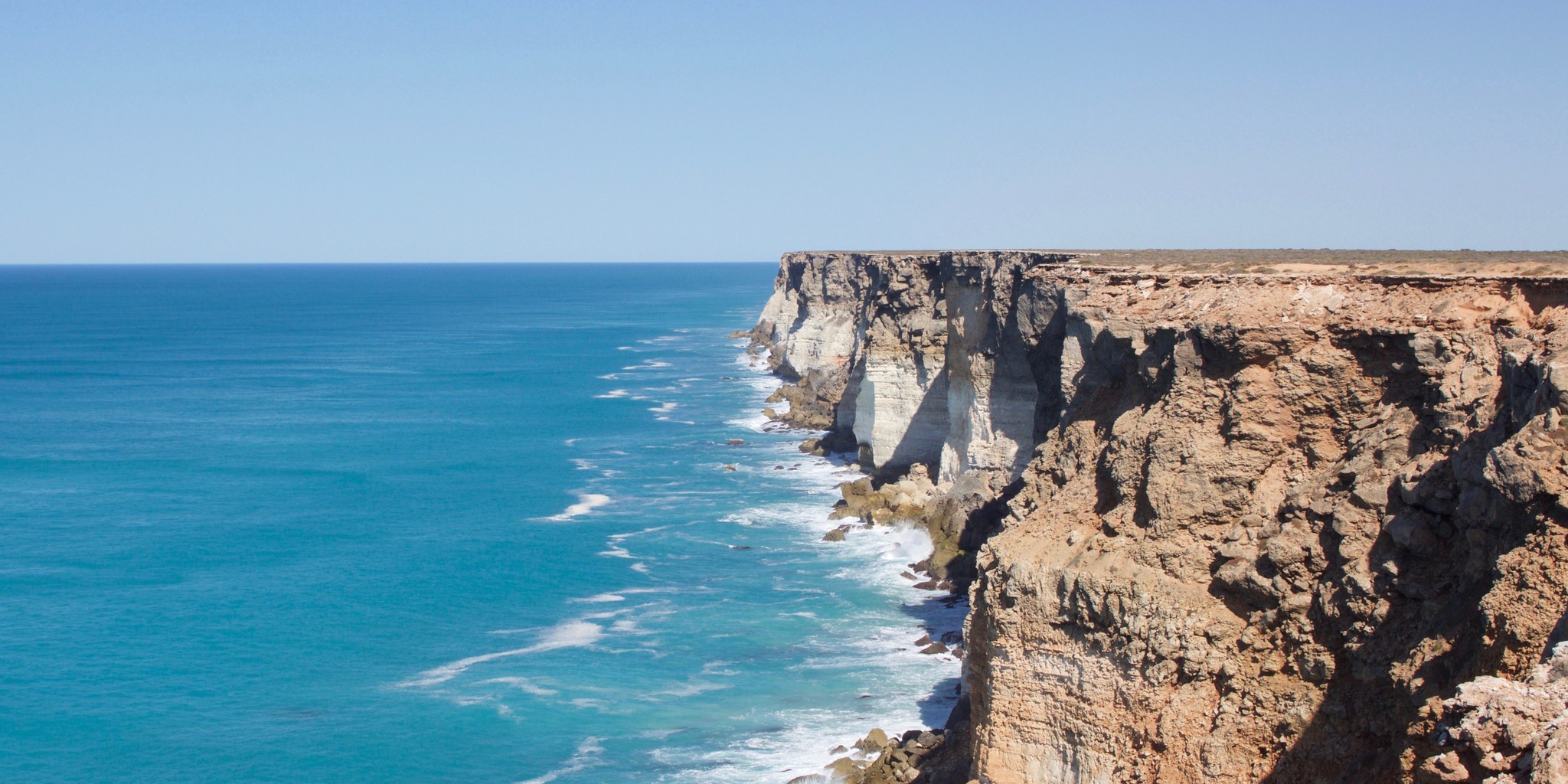 A photograph of the Great Australian Bight. To the right are steep rocky cliffs topped by a flat plain, and to the left is open water. The ocean is deep blue, growing darker towards the horizon. The sky is light blue and cloudless. Waves crash against the cliff face.