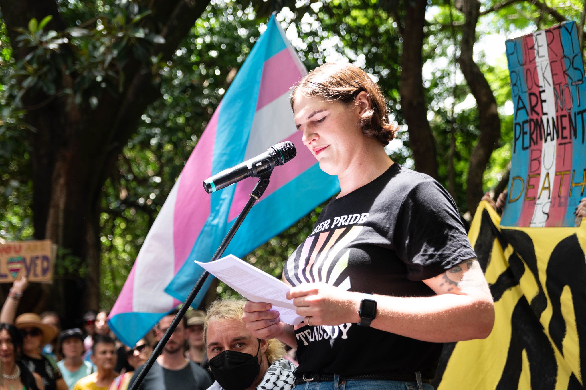 Necho speaking at the trans rights rally on 8 February in Magan-djin/Brisbane. A trans flag flies behind her and in frame is a sign that says "puberty blockers are not permanent but death is".