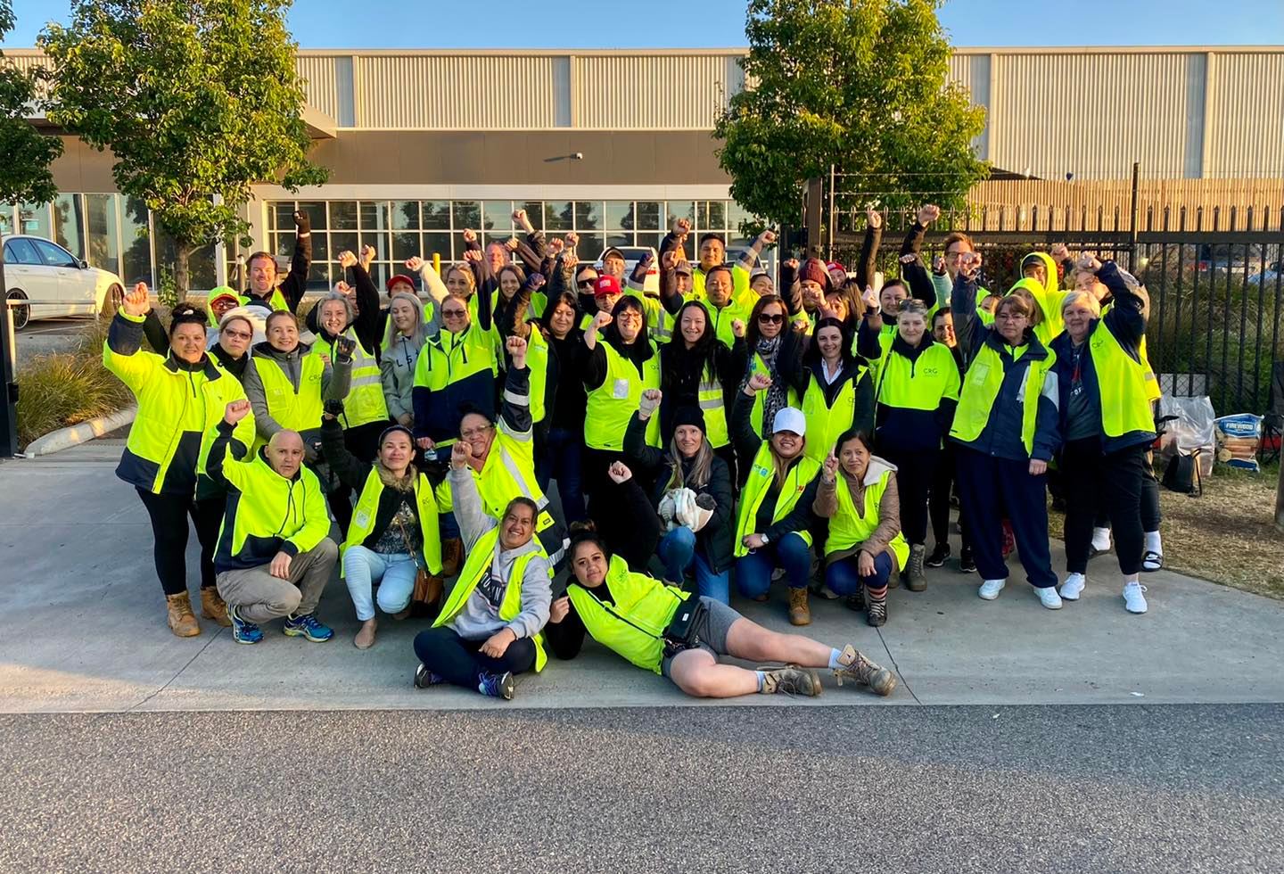  A photograph of a large group of Country Road workers wearing high-visibility fluorescent yellow jackets posing outside a warehouse with their fists raised in the air. Most of the workers are women. 