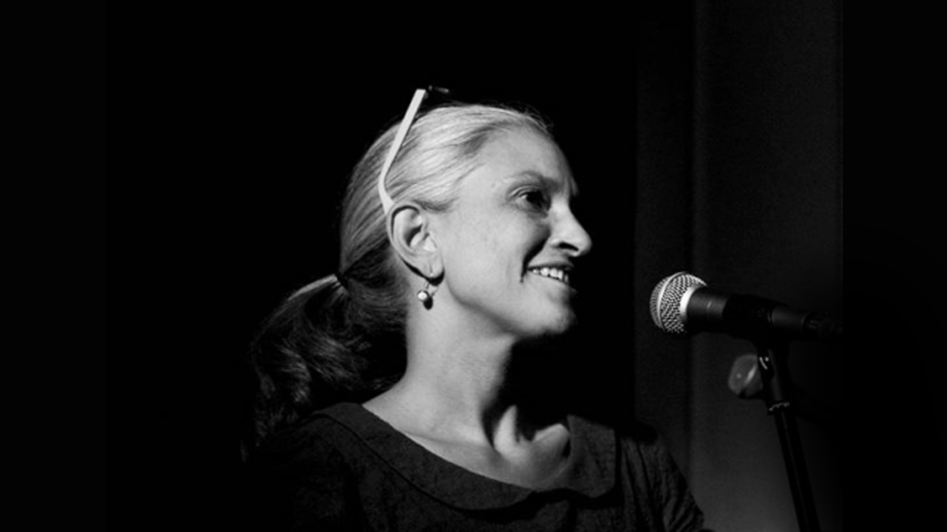 Black and white photograph of a woman speaking into a microphone. Only her face is clearly visible. She has fair hair and slender features.