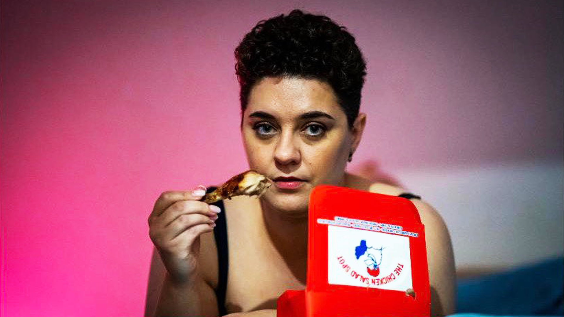 A young woman with short black hair, staring into the camera and holding a cooked chicken drumstick in their right hand. An open takeaway food box is nearby.