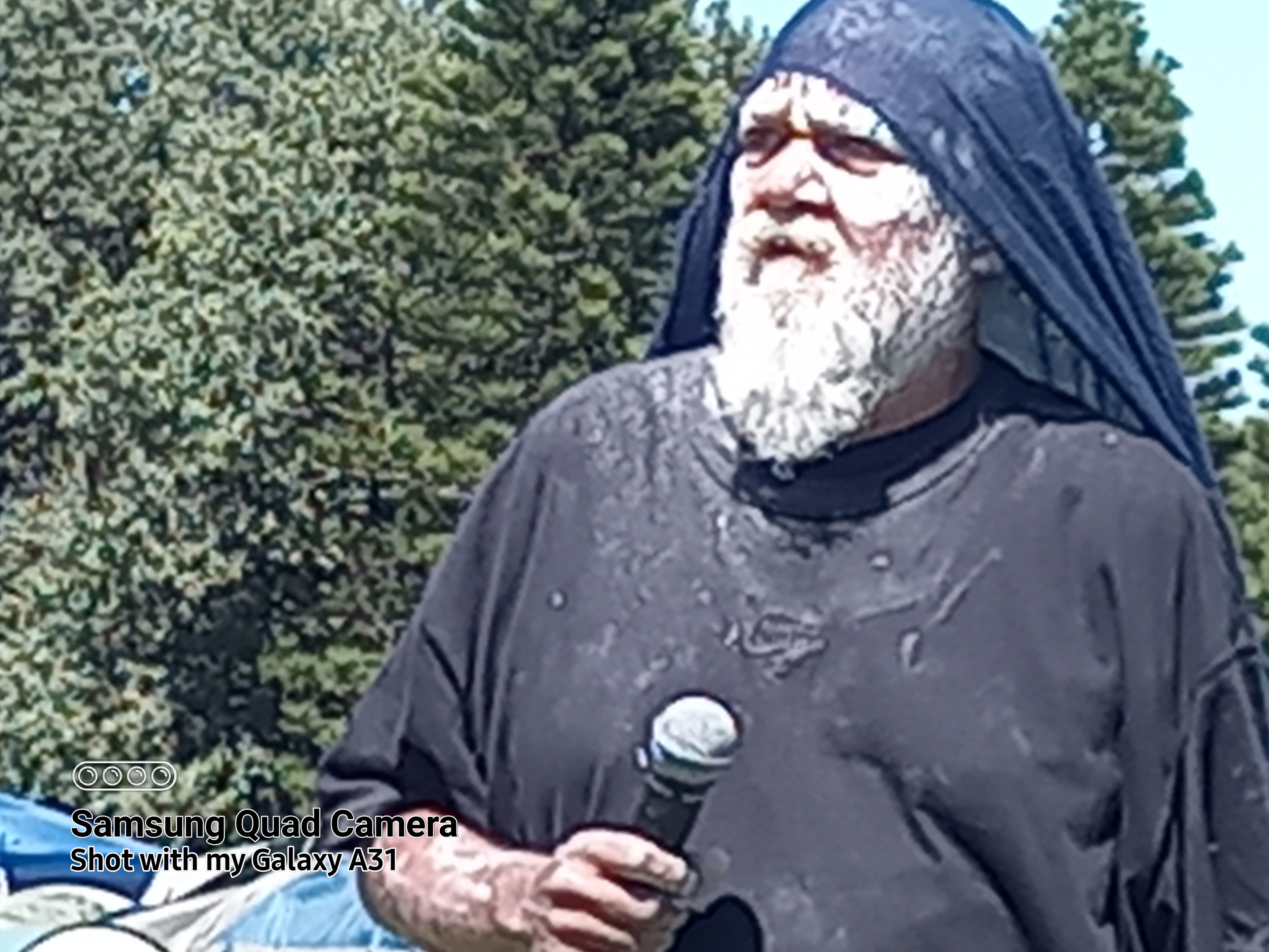 A First Nations bearded elder man, with white paint face, black clothes and microphone