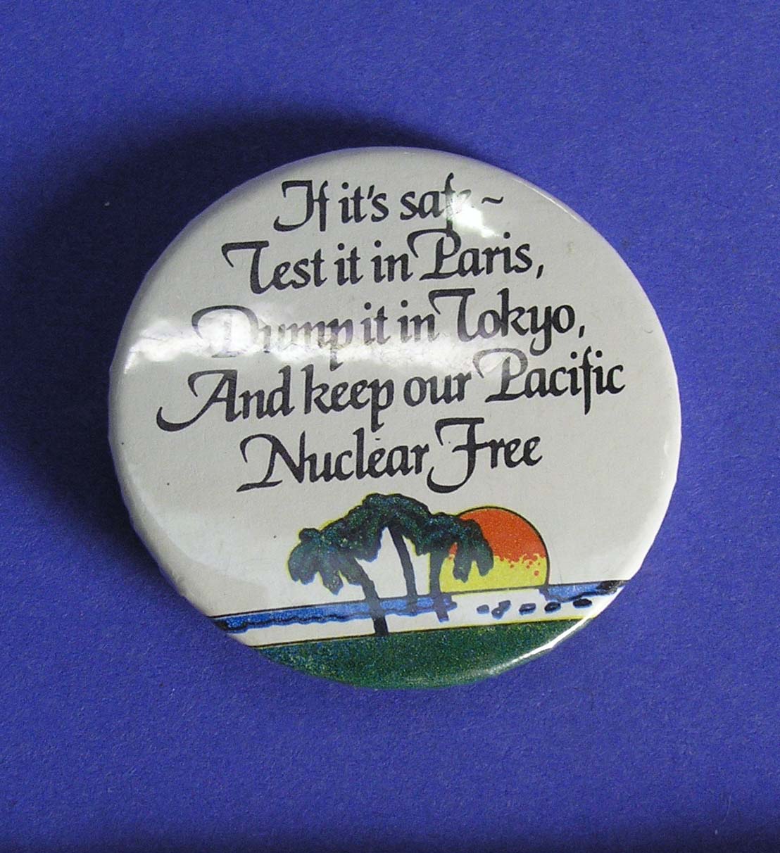  Anti-nuclear protest button badge; black lettering on white ground, "If it's safe - - Test it in Paris - Dump it in Tokyo - And keep our Pacific - Nuclear Free" above Pacific island scene of sun setting behind coconut palms