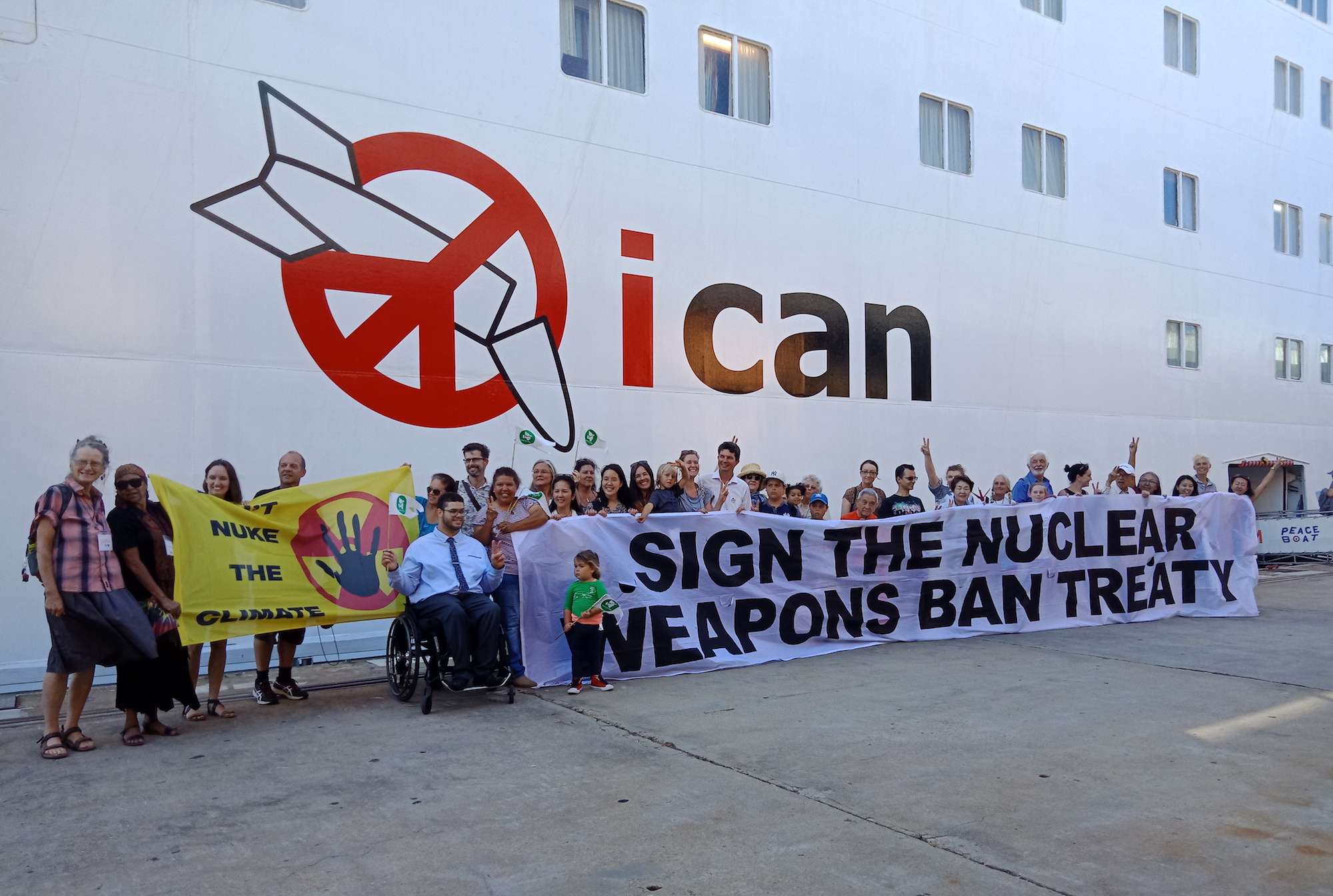 A large number of people holding two banners in front of a cruise ship - the peace boat - which has ICAN written on side with the ICAN logo. One yellow banner on left reads "Don't nuke the climate" and a larger white banner on the right reads "Sign the nuclear weapons ban treaty". Most people are standing behind the banners, there is one man in a wheel chair between the banners, and one child standing in front.