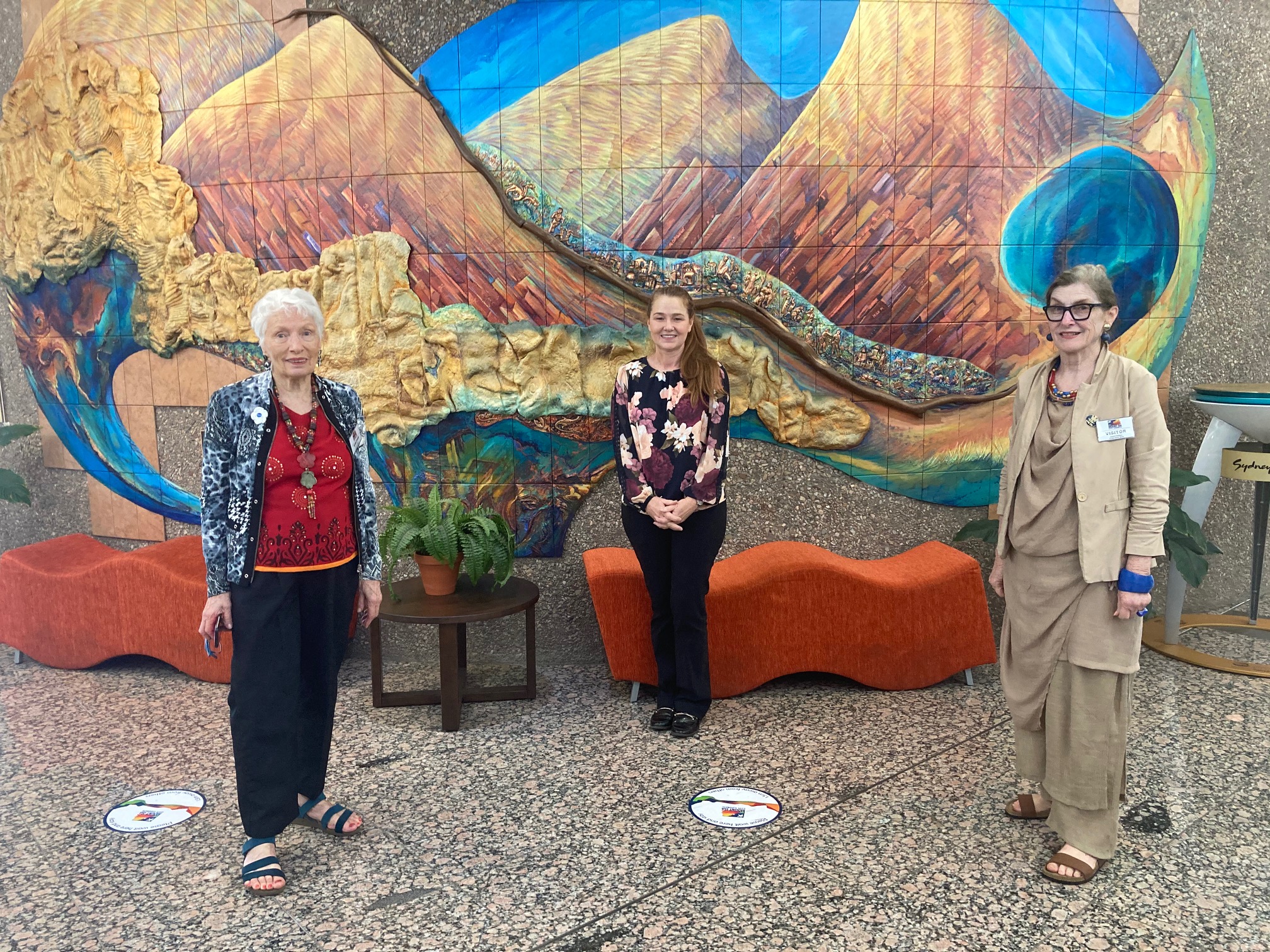 Three women, standing spaced out in front of a mural of mountains.