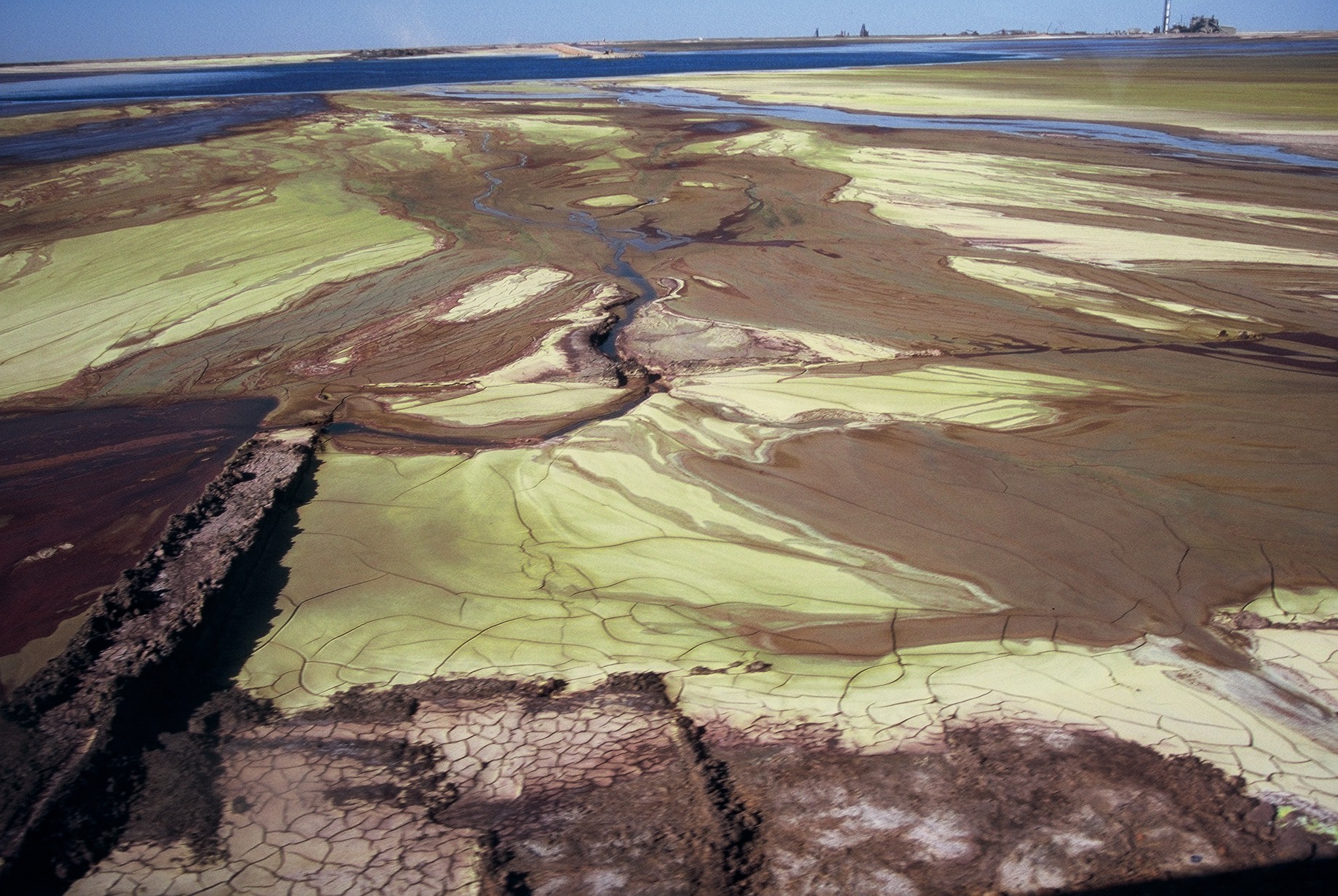 View of greenish-brown sludge-like ground, dry in some areas with small creeks. There is a road or path in the lower left corner.