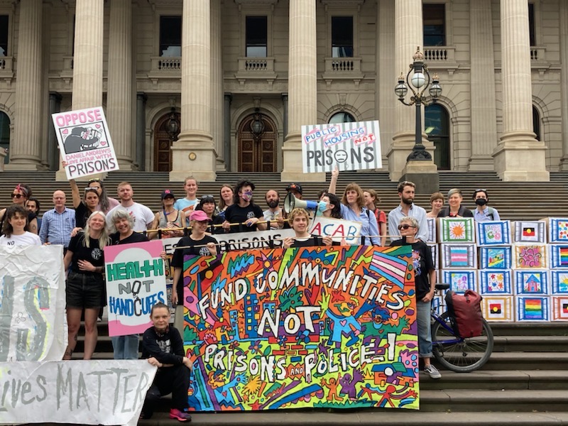 People gathering on the steps of Parliament in Naarm Melbourne with colourful banners says Homes Not Prisons or Police 