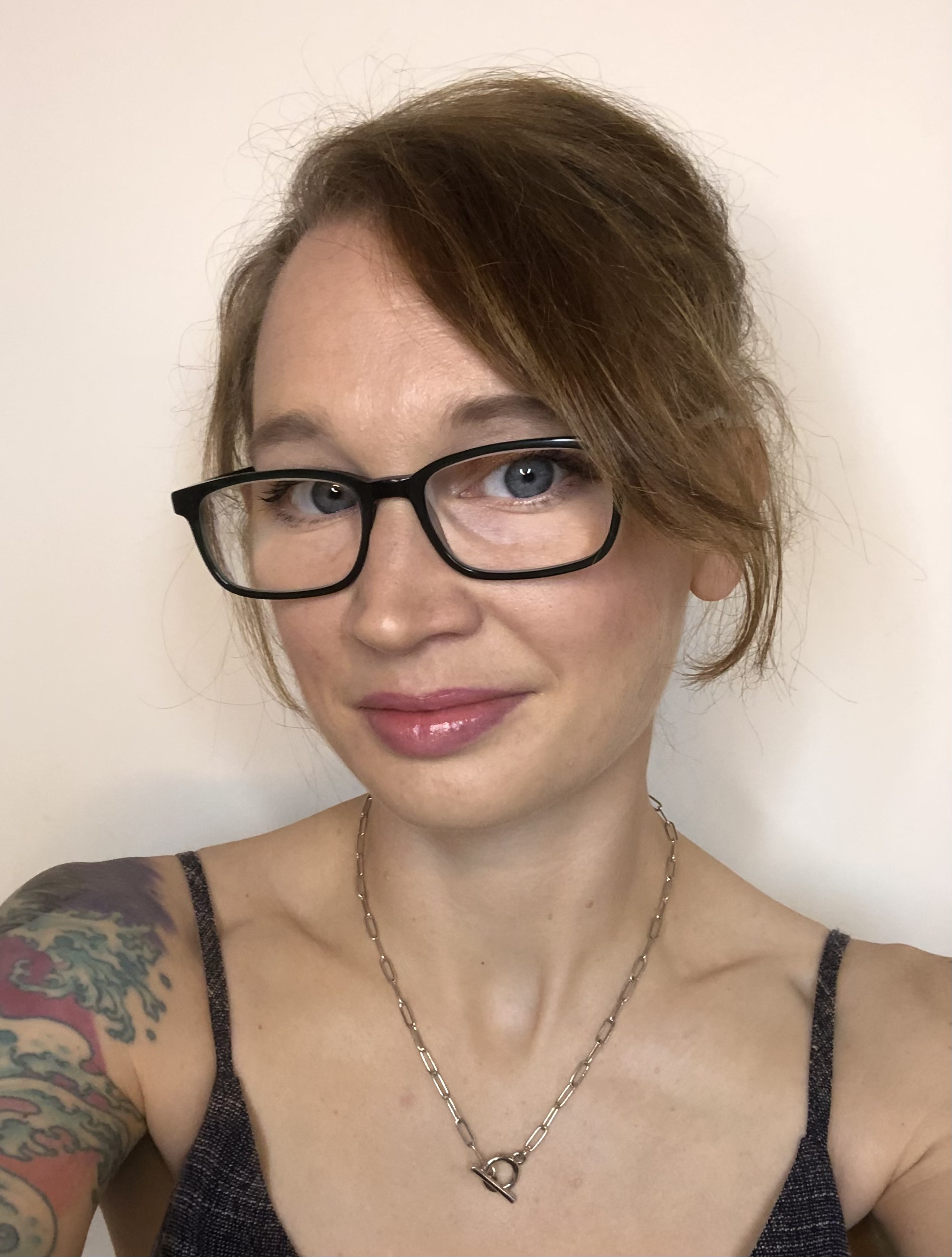 Photo portrait of a fair skinned woman with ligth brown hair, black rimmed glasses and tattoo on the right shoulder, wearing a singlet top and fine chain necklace.