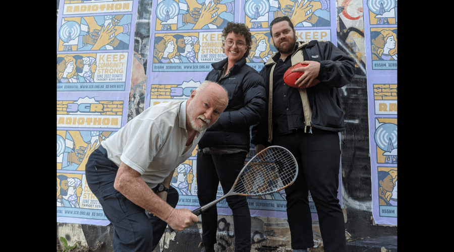 The sporting record hosts Em, John and James standing in front of a graffitied wall holding sports-related items