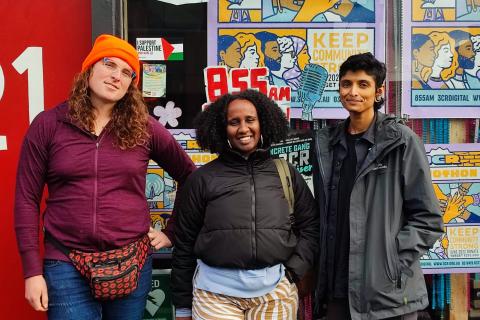 A photograph of Iris, Ayan and Priya standing in front of the 3CR station. The front window behind them is covered in 2022 Radiothon posters.