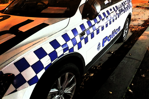 A white police car featuring the distinctive dark blue check pattern is parked next to a dark grey kerbside under orange street lights. Dark blue text on the side of the car reads Police