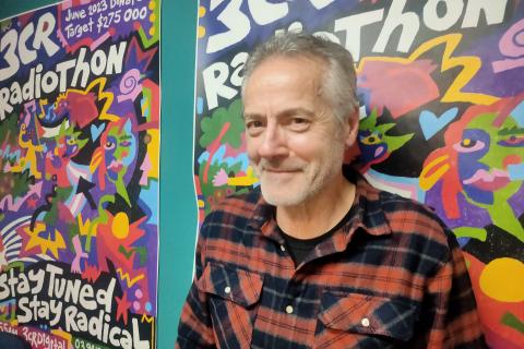 A man with greying hair wears a red and black check shirt. He looks to the side smiling