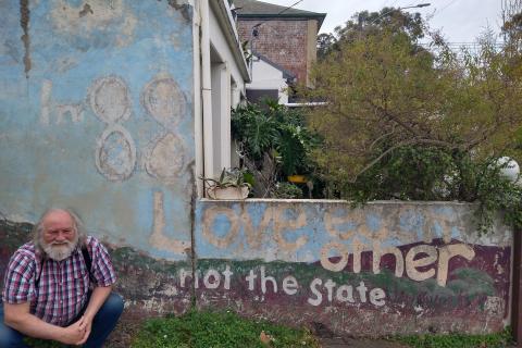 Don crouches down in front of a garden wall which reads 'In 88 love each other not the state'