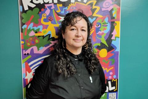 A woman with dark long curly hair and wearing a dark jumper looks sideways towards the viewer. She stands in front of a colourful poster