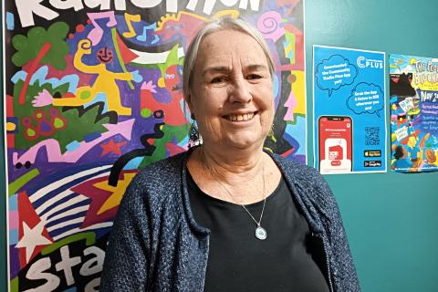 Sue looks towards the viewer wearing a black dress and blue cardigan. She stands in front of this year's colourful 3CR Radiothon poster and smiles 