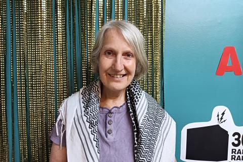 Sue stands in studio one at 3CR. Behind her is a gold sequined curtain. Sue wears a Palestinian kufiya around her shoulders.