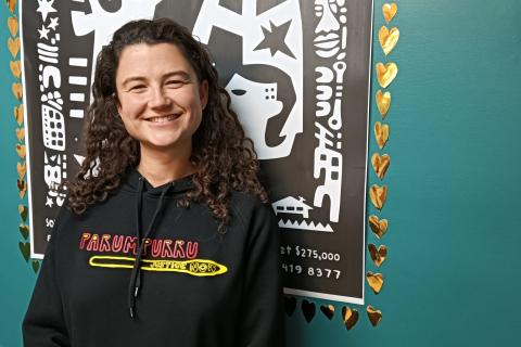 Rosie Heselev smiles for the camera in studio 1 at 3CR. She has shoelder-length curly brown hair and wears a black hoodie. She beams at the viewer.