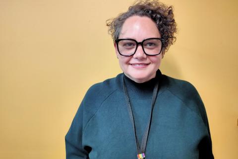 A woman in her forties looks into the camera. She wears a green jumper and black glasses. She has short curly hair