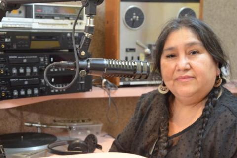 A woman sits in a radio studio among equipment. She wears a black top and has long black hair. She looks with a gentle smile towards the viewer