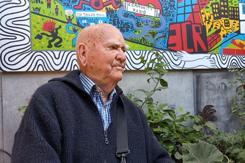 Percy sits in the 3CR courtyard and the colourful mural is behind him. He wears a blue, knitted jumper.