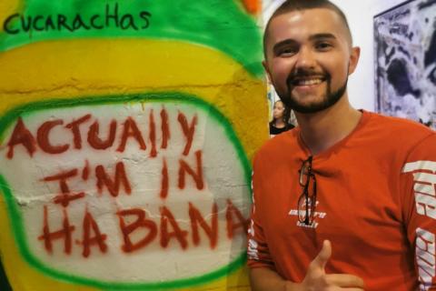 Sasha wears a red long-sleeve top and he stands next to a brightly-coloured pole with a big smile on his face