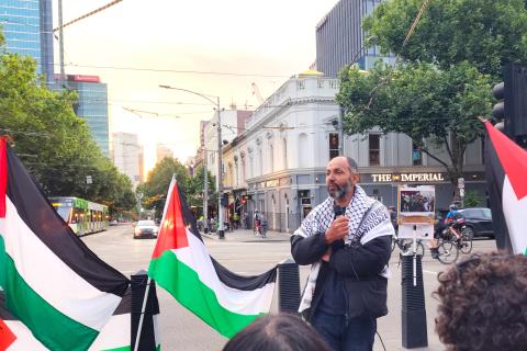 Uncle Ihab from Sit-Intifada on Wed 15 Nov 2023. Uncle Ihab is wearing black and white and black Keffiyeh holding a microphone next to three Palestinian flags, with trees, sunset and trams behind him.  