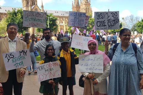 A group of racially diverse migrants hold signs in English and Arabic at a protest for climate justice.