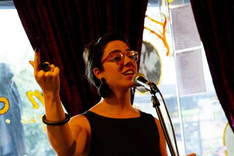 Ren Alessandra, young woman speaking live behind a microphone, one hand gesturing expressively