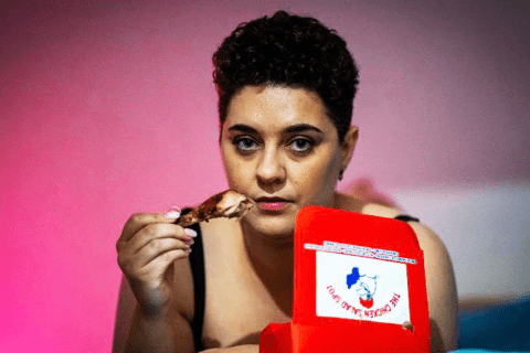 A young woman with short black hair, staring into the camera and holding a cooked chicken drumstick in their right hand. An open takeaway food box is nearby.