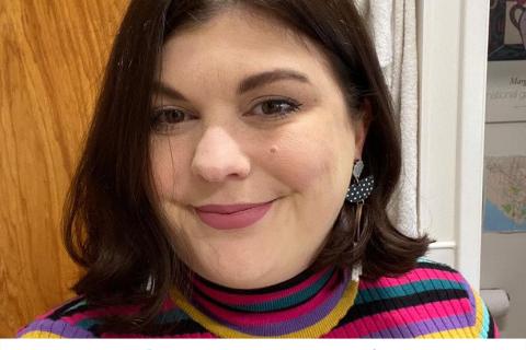 Steph Fornasier is a caucasian woman with shoulder length brown straight hair, wearing a pink, navy blue and gold striped skivvy and she is standing against a wooden back drop as she smiles at the camera.