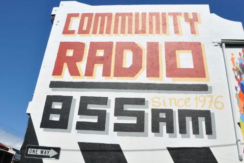 A photograph of the side of the 3CR station building in front of a clear sky. The text painted on the wall reads COMMUNITY RADIO- Since 1976 - 855 AM